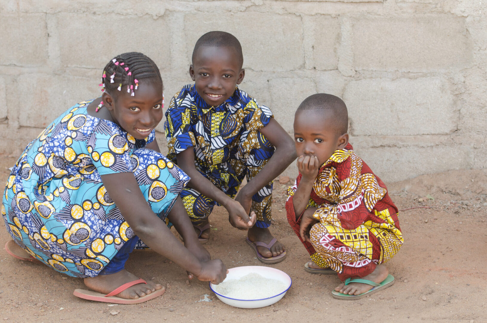 Children in colorful clothing eating together outdoors.
